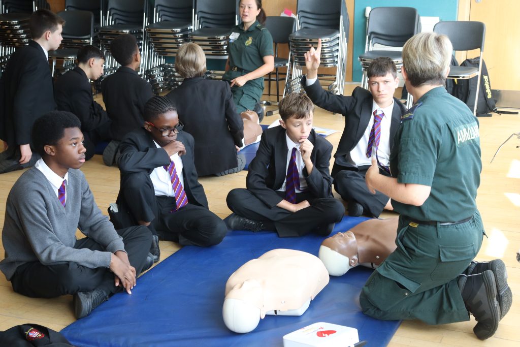 A London Lifesavers trainer leading a CPR training session with pupils. One pupil has his hand up to ask a question.