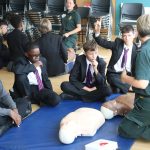 A London Lifesavers trainer leading a CPR training session with pupils. One pupil has his hand up to ask a question.