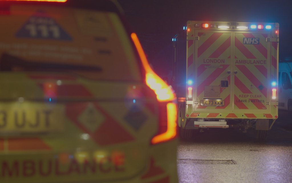 A close up of a fast-response car with an ambulance on blue lights in the background.